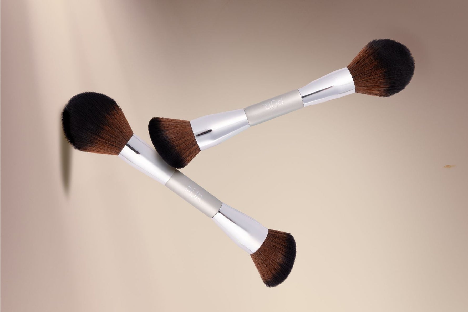 Two double-sided makeup brushes with silver handles and brown bristles arranged diagonally on a warm beige background.