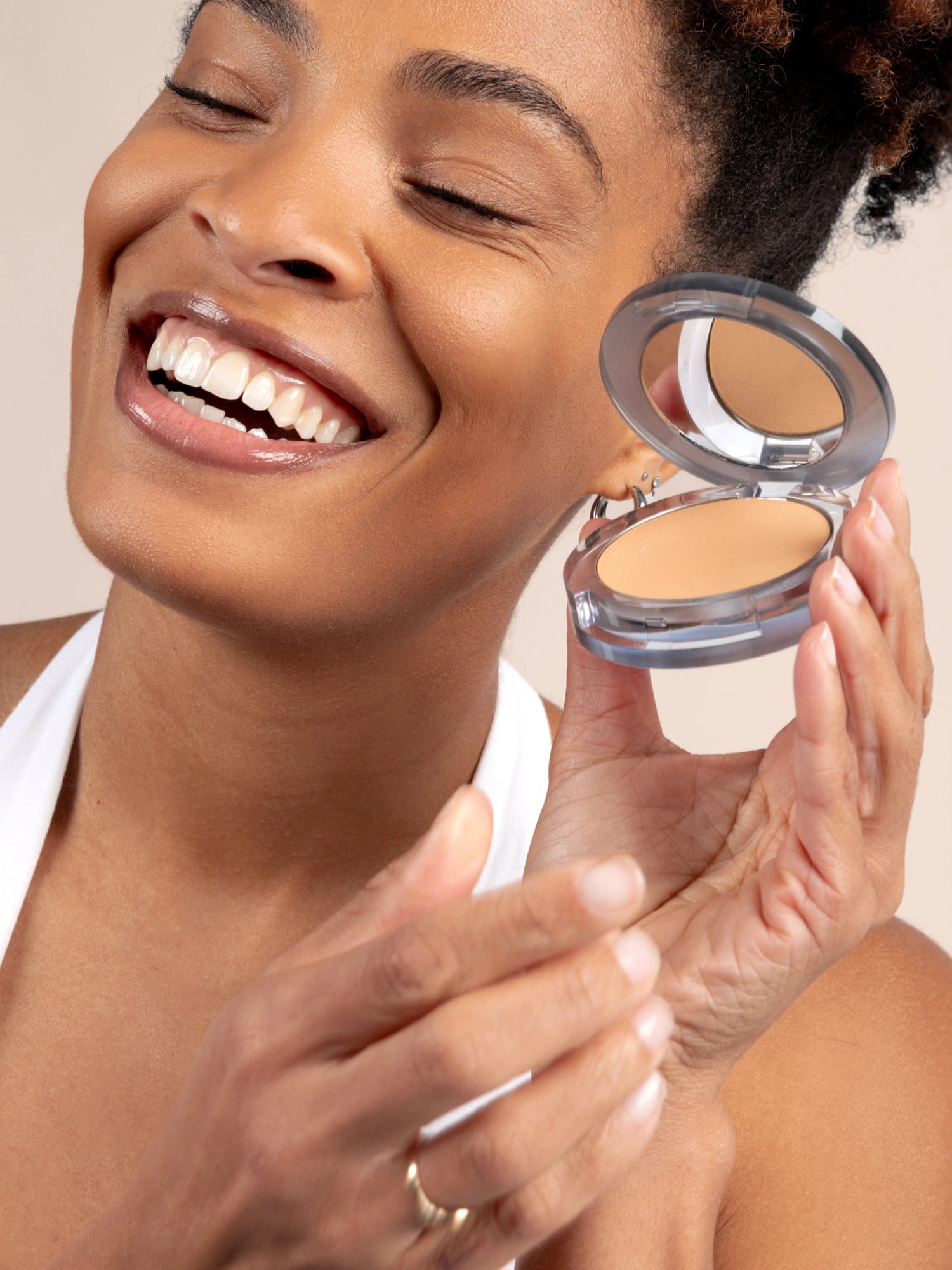 Model smiling with eyes closed holding 4-in-1 Pressed Powder against cheek - against a beige tan background