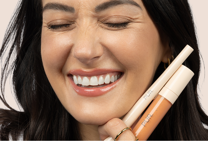 National Lipstick Day: 40% Off Lipstick - Woman smiling and holding Silky Sculpt and Glossy Lip Tint (Guava Glow) next to her face against pink background