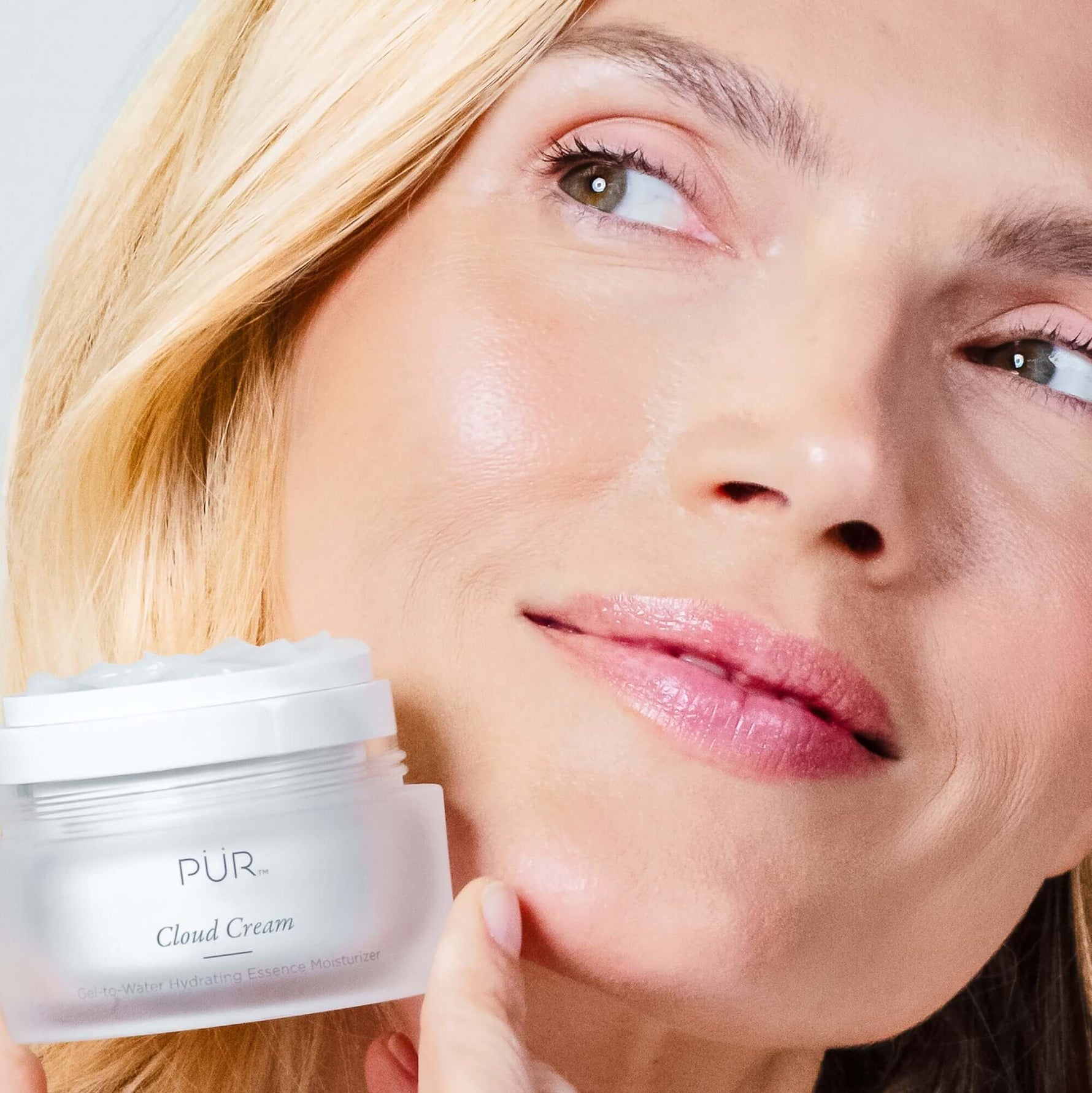 Close-up of a woman with glowing skin holding a jar of PÜR Cloud Cream Hydrating Essence Moisturizer.